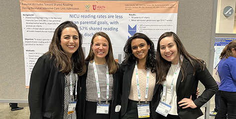 Four women smiling as they stand in front of a research poster at a conference.