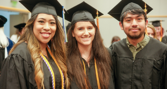 Three nursing students smile at the camera in graduation regalia