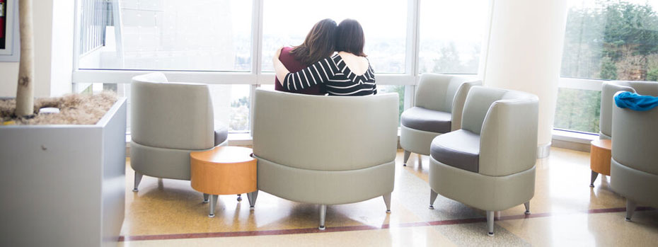 The backs of two women sitting next to each other and embracing in a hospital waiting room.