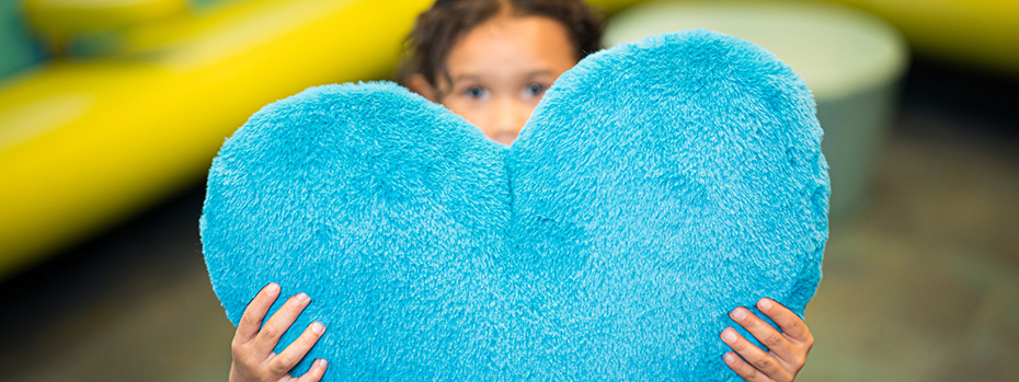 A young child holds up a blue heart-shaped pillow with both hands.