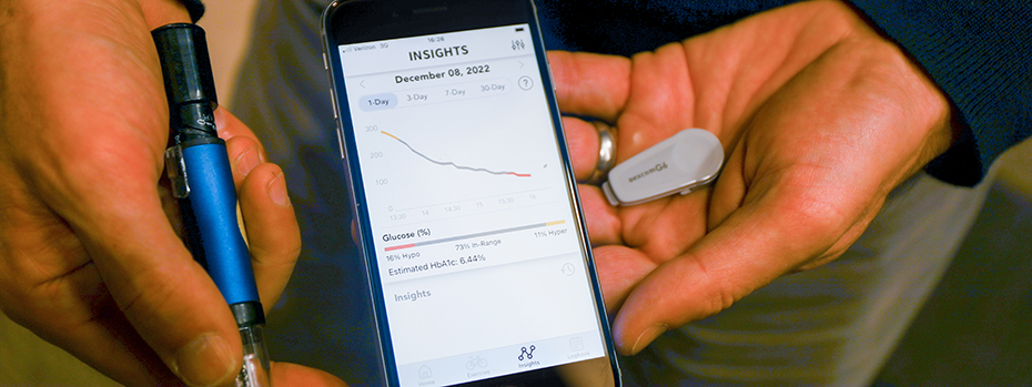 Hands holding glucose sensor transmitter, a smart insulin pen and phone displaying DailyDose app.
