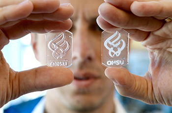 Hands holding up two small glass plates with opaque, engineered material in the OHSU torch logo. The researcher's face is in the background.