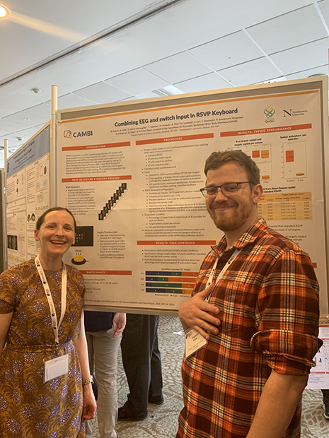 A woman and man stand smiling in front of research poster at the International BCI Meeting in Brussels.