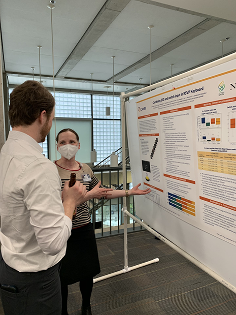 A woman wearing a PPE face mask discusses her research poster with a colleague.