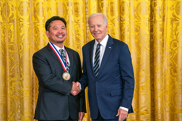 Dr. David Huang and President Joe Biden shake hands in front of a gold background.