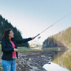 A woman is fishing on a verdant riverbank. She casts the rod with outstretched hand. The scene is peaceful and calm.