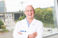 A doctor smiling with his arms crossed in front of a window through which Portland's South Waterfront is visible.