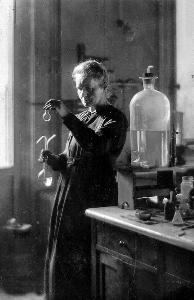Marie Curie stands in a long black dress in her laboratory, looking into glass test tubes.