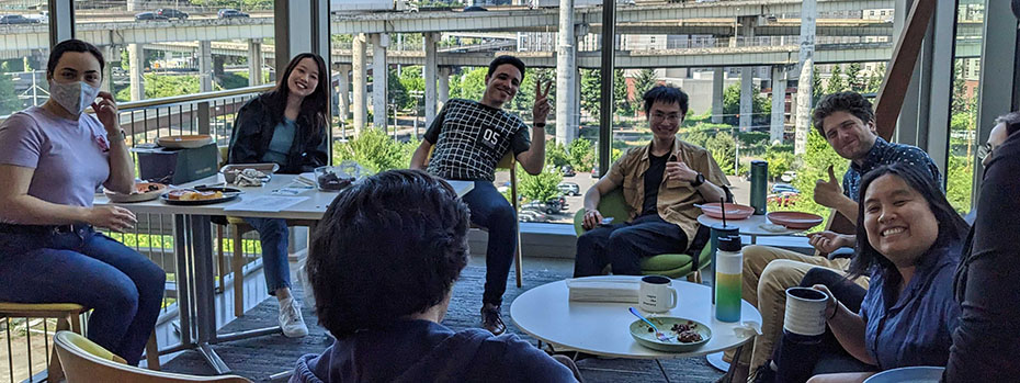Nine students gather around two tables with plates of food and beverages. They are smiling and gesturing to the camera. Overpasses and bridges are visible through large glass windows in the background.