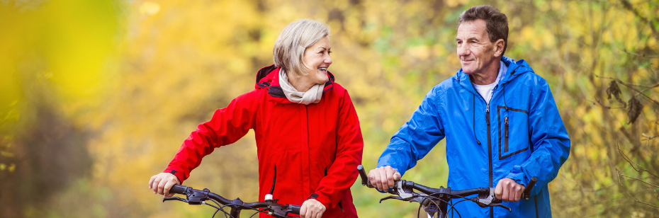 Two older adults walking with their bicycles near colorful fall bushes
