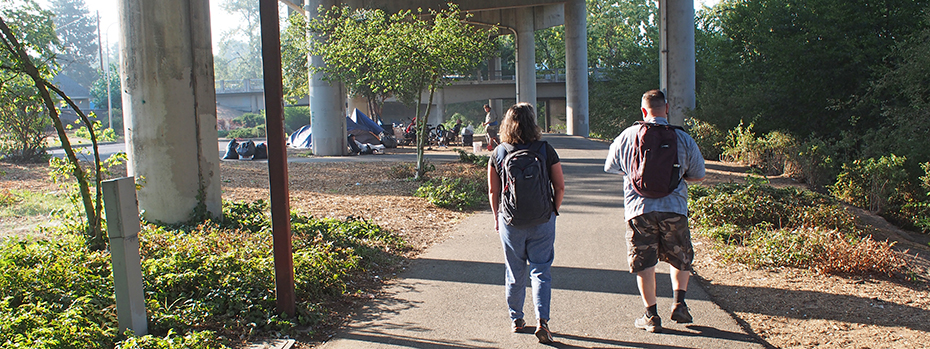 Two community members walk under a bridge. A group of tents and unhoused people are nearby.