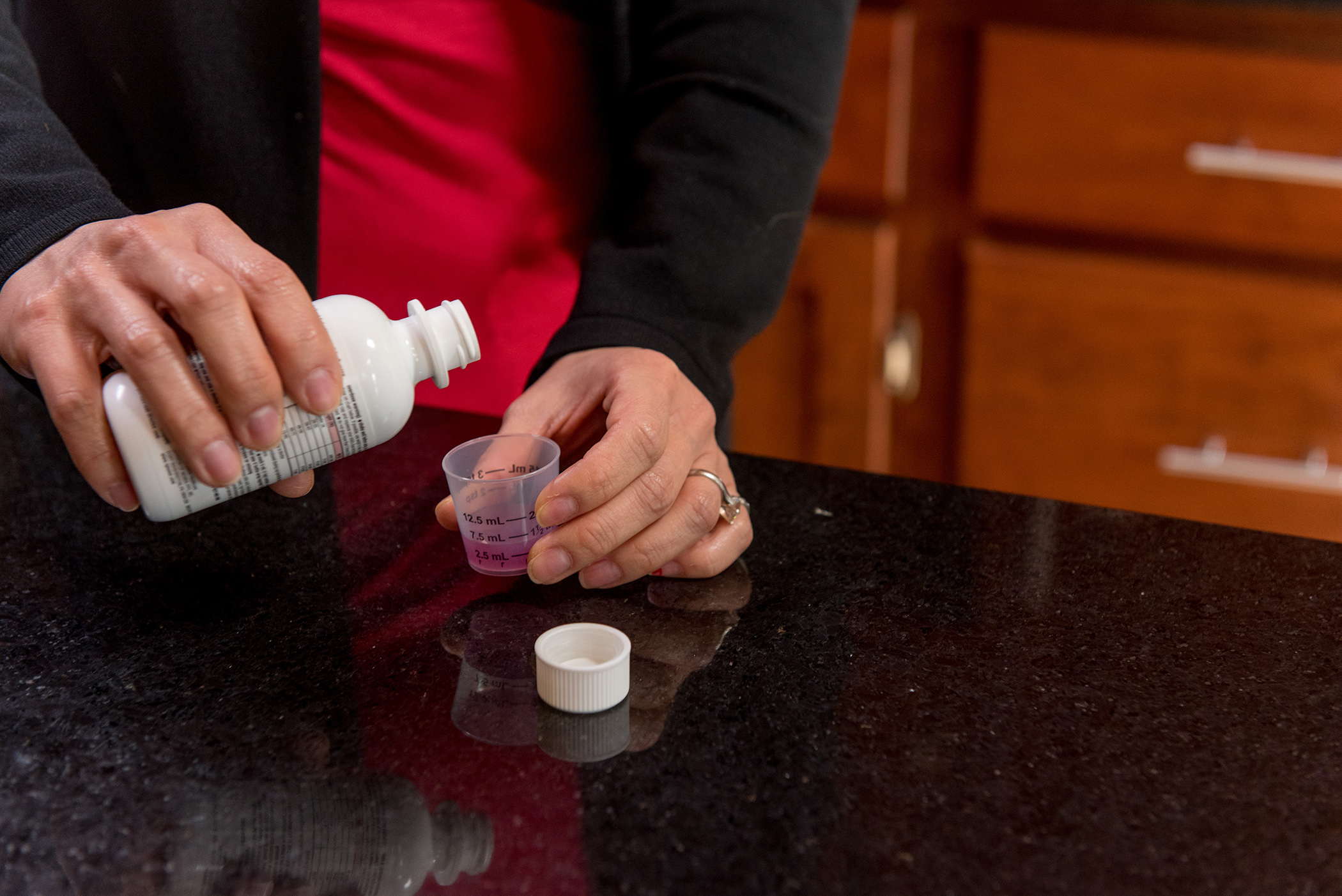 A person pours pink liquid medicine from a white bottle into a dosing cup over a black kitchen counter