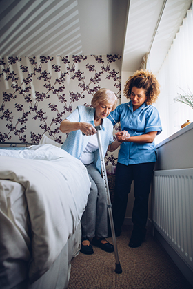 Home Caregiver helping a senior woman get up from her bed with a walking stick.