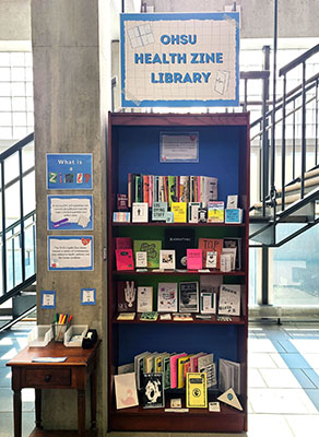 Tall shelf with sign "OHSU Health Zine Library" stands in front of BICC Library stairs, filled with colorful zines