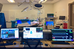 Three computer monitors set up outside of a simulation lab operating room window.