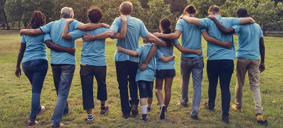 9 people walking away, side-by-side with their arms around each other in a park. They are all wearing the same color t-shirt.