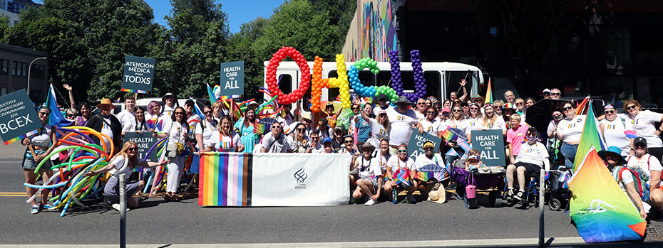 OHSU employees pose for a large group photo with balloons that spell OHSU and an OHSU banner with stripes in LGBTQ+ colors. 