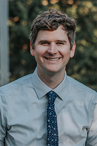 SON professor Andrew McHill smiles at the camera while standing in front of green bushes.