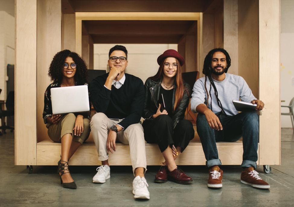 A diverse group of four post docs and early career faculty sit on a wooden bench holding laptops and books and looking happy to be there together.