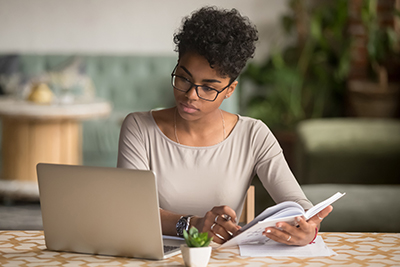 Focused young african american businesswoman or student looking at laptop holding book learning