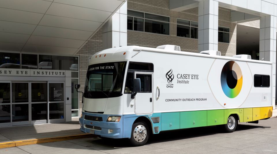The mobile clinic is a large RV with the Casey Eye Institute logo. it's parked in front of the Casey Eye Institute building. 