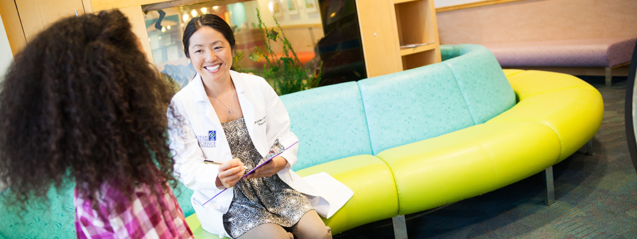 Christine Sayama, an OHSU pediatric neurosurgeon, holds a pen and clipboard while sitting with a young person in a waiting area at OHSU Doernbecher Children’s Hospital.