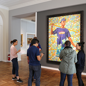 Four students stand in a semicircle in front of a painting depicting a young man in a blue T-shirt against a floral background.