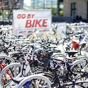 Rows of parked bicycles sit in front of a white sign with red letters that read, “Go By Bike.”
