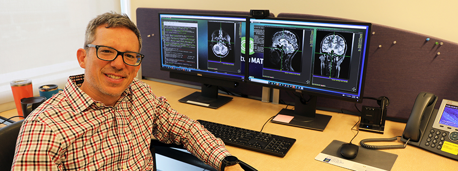Juan Piantino, an OHSU pediatric neurologist, sits in front of two computer monitors showing images of brains.