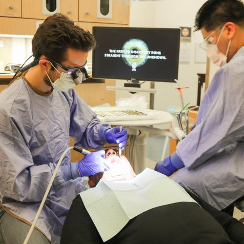 A dentist uses 2 instruments in each hand to perform a dental procedure on a patient. A dental hygienist sits on the other side of the patient observing.