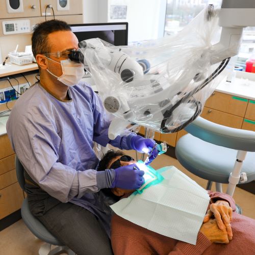 A dentist works in a patient's mouth while looking into a magnifying device suspended from above. The patient lies calmly during their procedure with their hands clasped on their stomach.