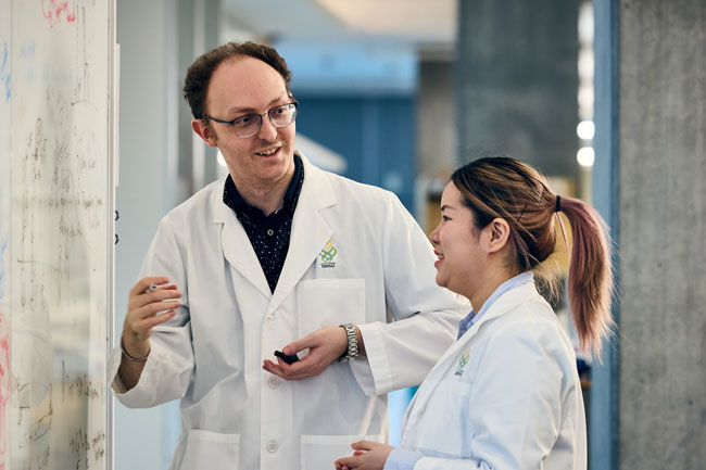 Two researchers with white coats on standing in a lab having a discussion.