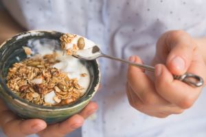A person in a white shirt eats a healthy meal of yogurt and granola with pumpkin seeds.