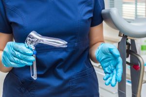 A provider in blue scrubs and gloves holds a plastic speculum in their hands.