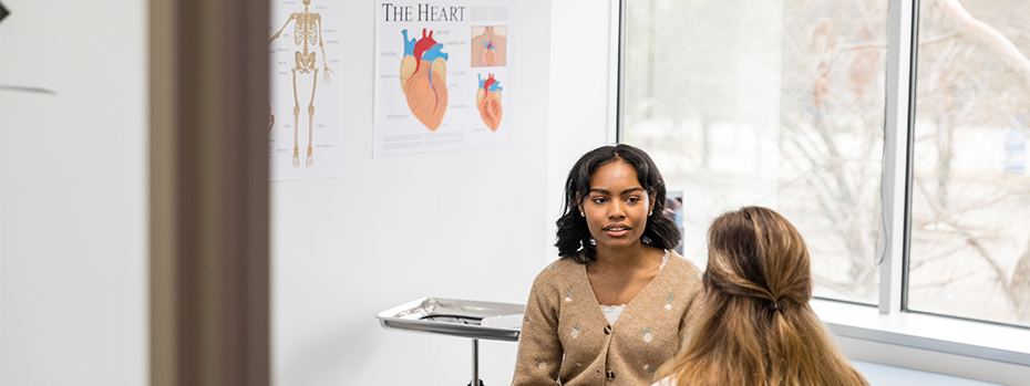 A young female patient sits on an exam table talking to a female doctor.