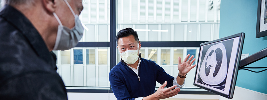 A doctor gestures to an image of lungs on a computer monitor. A patient wearing a face mask sits nearby.