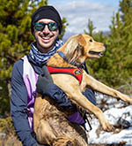 A man wearing sunglasses and outdoor gear holds his dog while standing in front of a snowy field.