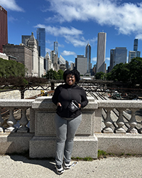 A woman standing on an overpass above a highway on a sunny day with a city in the background.