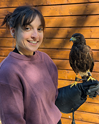 A woman smiling wearing a protective glove on her hand which has a hawk perched on it.