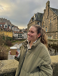 A woman standing on a bridge smiling holding a coffee with a town visible in the background.