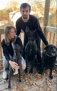 A man and a woman on a porch crouch down behind three black dogs on a sunny autumn day.