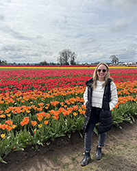 A woman standing outside in front of a flower field smiling.