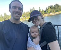 A man takes a selfie next to a woman holding their baby outside in front of a lake.