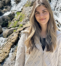 A woman smiling outside while on a hike with a rocky slope behind her.