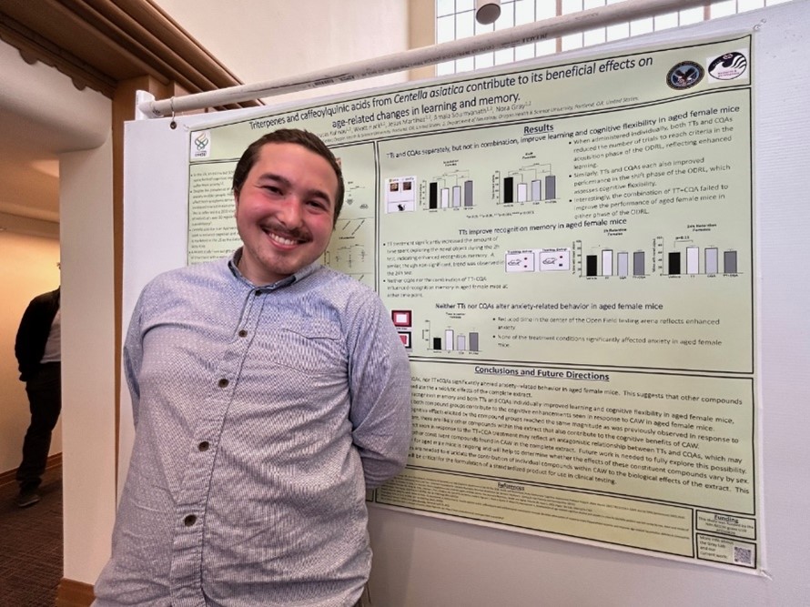 A man wearing a light blue button-down shirt stands smiling in front of his scientific poster at a conference.