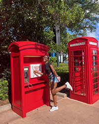 A woman smiling while posing in front of a red payphone on a sunny day.