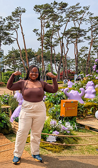 A woman standing and smiling as she points behind her to a forest park on a sunny day.