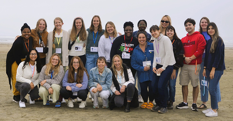 A group of twenty people standing and crouching together smiling at the beach on a cloudy day.