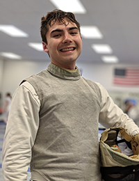 A man in fencing attire holding his mask under one arm smiling.