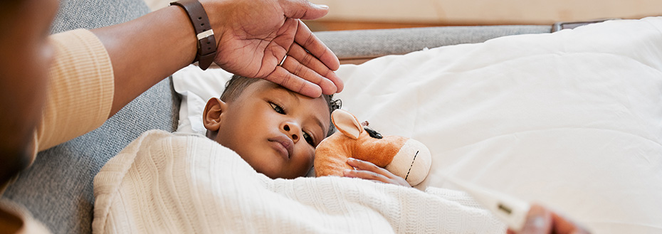 A young child snuggles a stuffed animal in bed while his parent feels his forehead and holds a thermometer.